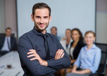 Portrait of successful business leader standing with crossed arms and smiling, business people sitting at office desk on background
