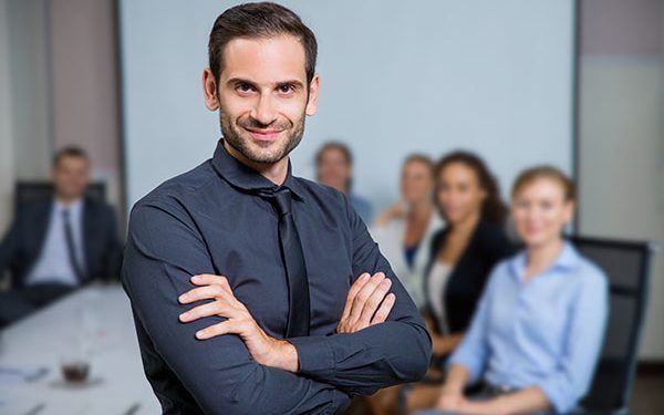 Portrait of successful business leader standing with crossed arms and smiling, business people sitting at office desk on background