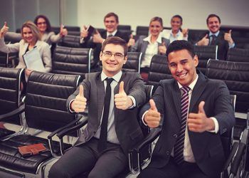 Two happy young businessmen showing thumbs up on background of their colleagues