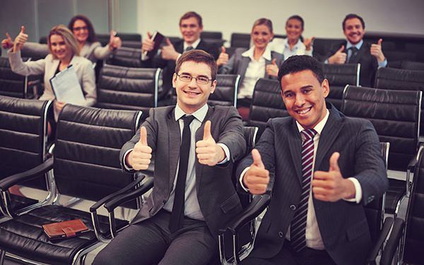 Two happy young businessmen showing thumbs up on background of their colleagues