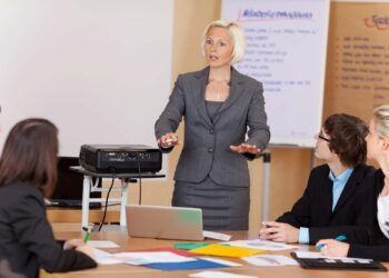 Woman sung a projector giving a corporate training class to a group of young businesspeople around a table gesturing with her hands as she explains something