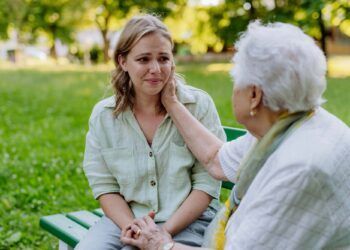 A worried senior grandmother comforting grown up granddaughter when sitting on bench in park, share problem with someone close concept