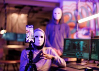 Mobile phone screen displaying anonymous hacker recording scam message. Internet criminals in masks and hoods live streaming on smartphone in abandoned warehouse at night