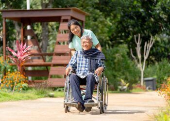 Asian nurse cart of shove senior man sit on wheelchair to walk along the walkway in the garden and they look happiness outdoor to relax with warm light.