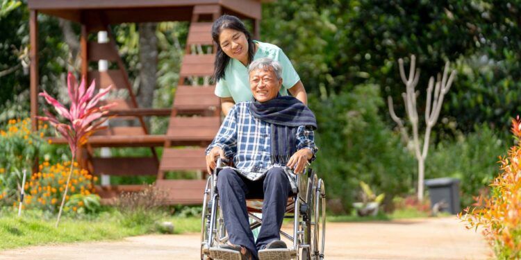 Asian nurse cart of shove senior man sit on wheelchair to walk along the walkway in the garden and they look happiness outdoor to relax with warm light.