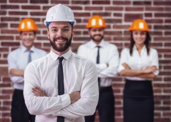 Handsome young bearded businessman in suit and protective helmet is looking at camera and smiling, his colleagues in the background