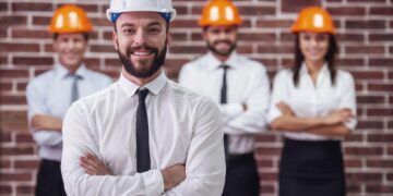 Handsome young bearded businessman in suit and protective helmet is looking at camera and smiling, his colleagues in the background