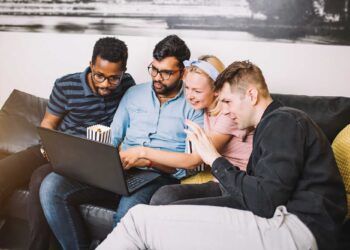 Group of young multi ethnic hipster best friends with laptop in home interior. Concept of friendship, fun, technology, home party. Young people laughing using computer together at home.