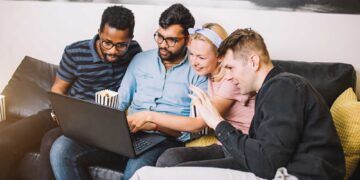 Group of young multi ethnic hipster best friends with laptop in home interior. Concept of friendship, fun, technology, home party. Young people laughing using computer together at home.
