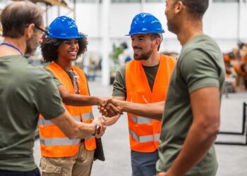 Multiracial male and female engineers wearing safety vests and hardhats shaking hands in a robotics factory, celebrating successful teamwork and collaboration in advanced technology development