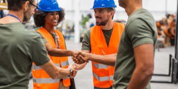Multiracial male and female engineers wearing safety vests and hardhats shaking hands in a robotics factory, celebrating successful teamwork and collaboration in advanced technology development